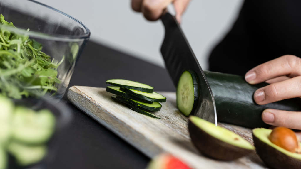 Bee Fit Coach, Shelby slicing a cucumber into round slices on a wooden cutting board.