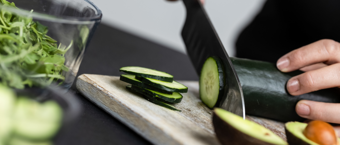 Bee Fit Coach, Shelby slicing a cucumber into round slices on a wooden cutting board.