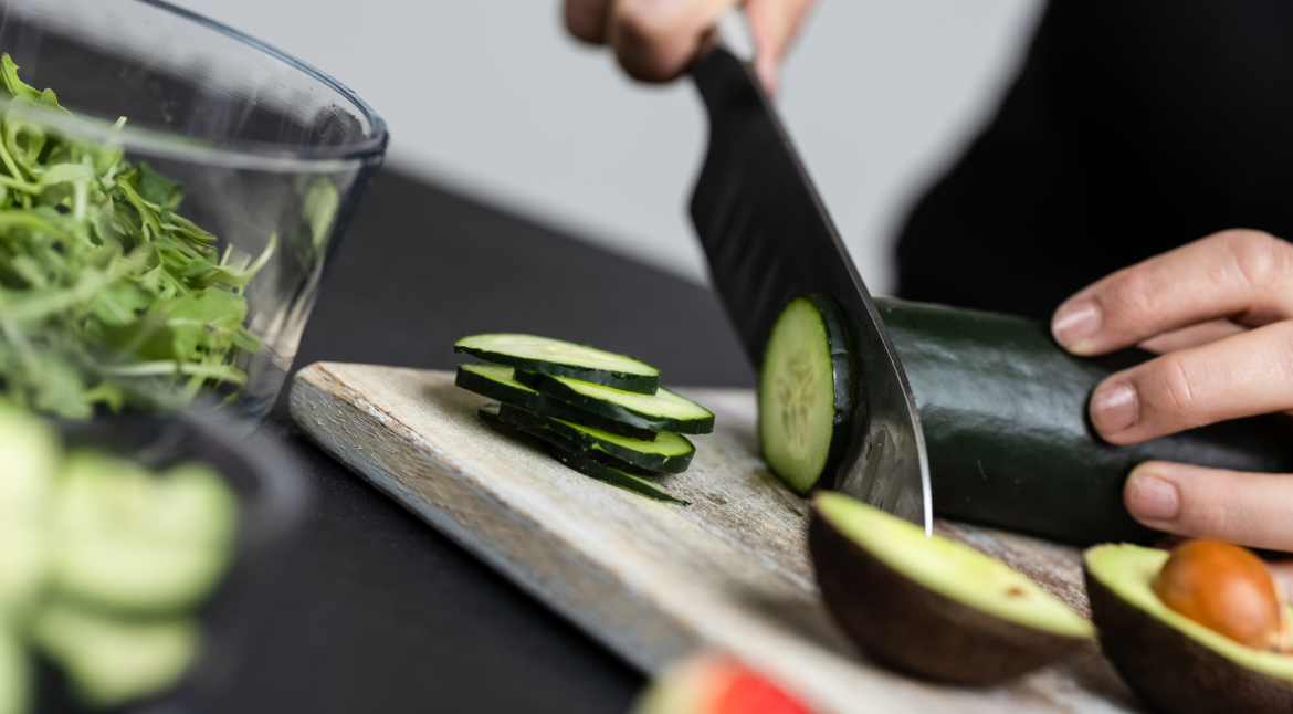 Bee Fit Coach, Shelby slicing a cucumber into round slices on a wooden cutting board.