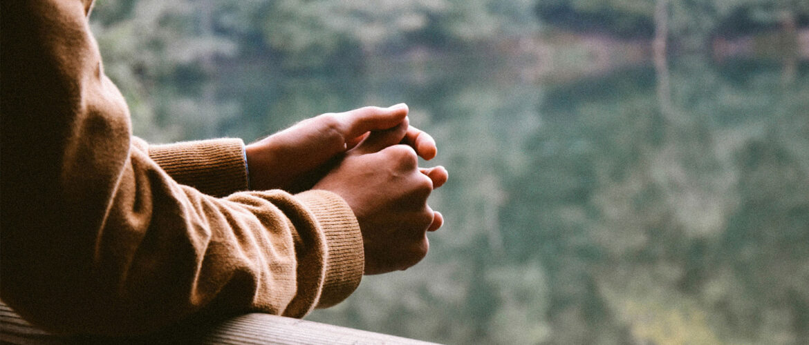 A person rests their arms against a railing overlooking a lake, and forest. They look to be in thought, and the photo is cropped at their shoulders. Original photo by Umit Bulut.