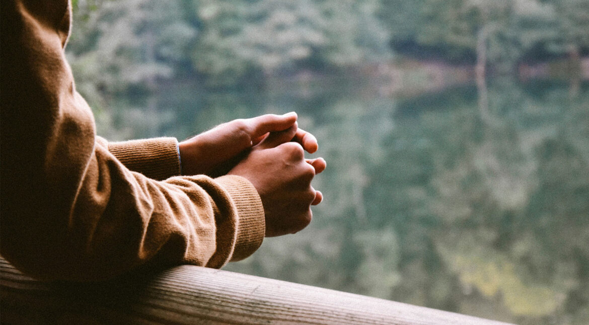 A person rests their arms against a railing overlooking a lake, and forest. They look to be in thought, and the photo is cropped at their shoulders. Original photo by Umit Bulut.