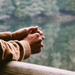 A person rests their arms against a railing overlooking a lake, and forest. They look to be in thought, and the photo is cropped at their shoulders. Original photo by Umit Bulut.